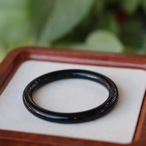 May include: A smooth, circular black bangle bracelet with a polished, reflective surface. The solid black band features a uniform, rounded profile and a glossy finish, presented as a single continuous loop of dark material without any additional ornamentation.