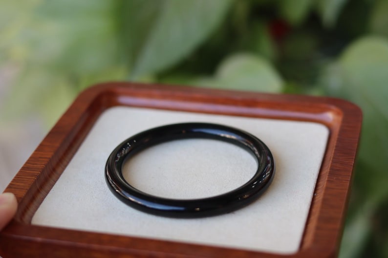 May include: A smooth, circular black bangle bracelet with a polished, reflective surface. The solid black band features a uniform, rounded profile and a glossy finish, presented as a single continuous loop of dark material without any additional ornamentation.