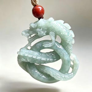 May include: A pale green carved jade pendant in the shape of a coiled dragon with intricate scale details. The pendant is suspended from a cord featuring a single, smooth, round, dark red bead at the top, providing a contrast in color.