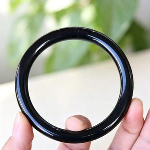 May include: A smooth, solid black bangle bracelet with a polished, reflective surface. The circular accessory features a rounded profile and a continuous, seamless design, showcasing a deep, uniform black color throughout the entire piece.