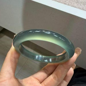 Può includere: Un braccialetto bangle in giada traslucida. Il braccialetto è una fascia liscia e arrotondata con una sfumatura di colori, che passa da un verde-azzurro intenso a un verde pallido. La superficie lucida riflette la luce, evidenziando le variazioni naturali della pietra.
