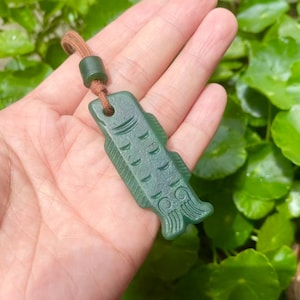 May include: A dark green carved stone pendant in the shape of a fish, featuring etched scale and fin details. The pendant is attached to a brown cord with a matching green cylindrical bead, creating a decorative accessory for a necklace or charm.