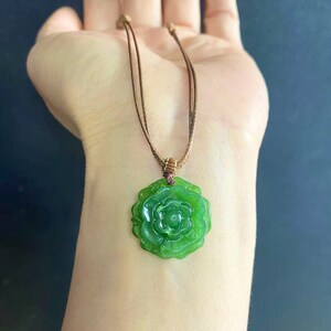 May include: A green jade pendant necklace featuring a carved rose design. The pendant is suspended from a brown cord necklace. The jade has a translucent quality, and the rose has detailed petals.