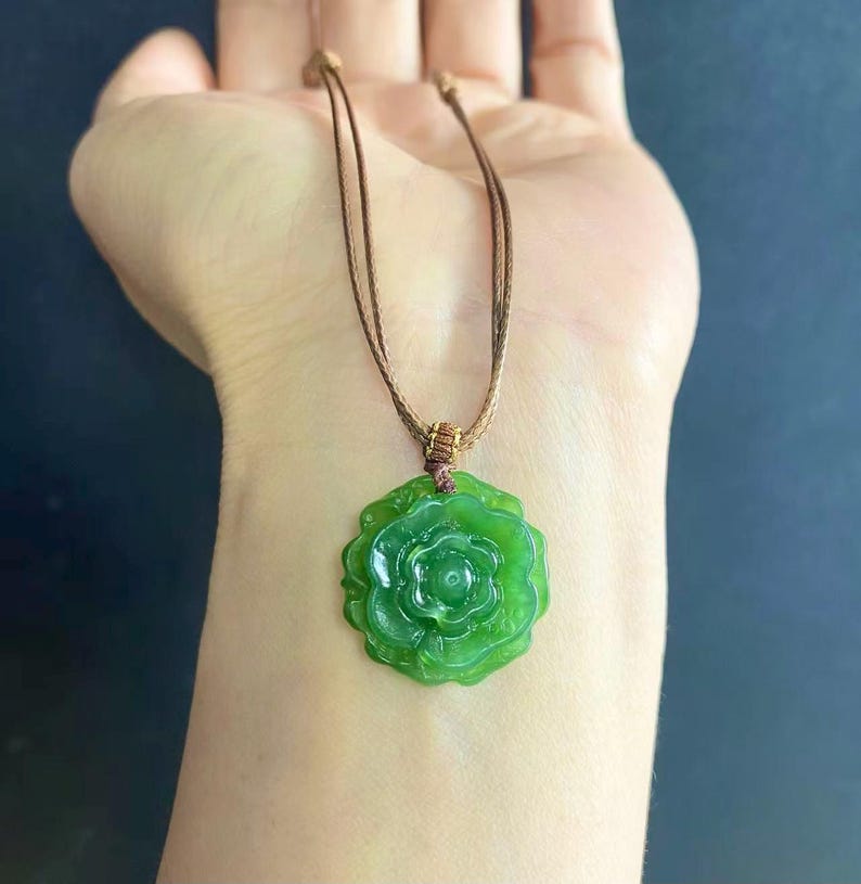 May include: A green jade pendant necklace featuring a carved rose design. The pendant is suspended from a brown cord necklace. The jade has a translucent quality, and the rose has detailed petals.