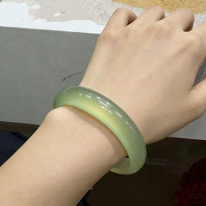 May include: A light green jade bangle bracelet. The smooth, polished surface reflects light, showcasing a translucent quality. The bracelet is a solid, circular design, and appears to be a natural stone. The color is a soft, pale green.