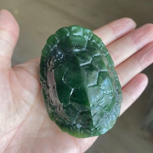 Pode incluir: Uma escultura de pedra verde-escura polida em forma de casco de tartaruga. A superfície apresenta uma textura lisa e brilhante com padrões distintos de placas hexagonais e pentagonais em relevo que imitam o aspeto natural do casco.