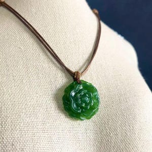 May include: A green jade pendant necklace featuring a carved rose design. The pendant is suspended from a brown cord necklace. The necklace is displayed on a mannequin.