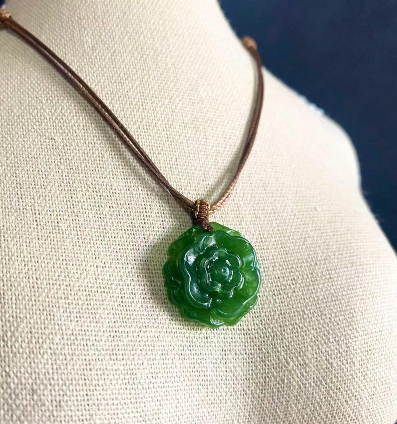May include: A green jade pendant necklace featuring a carved rose design. The pendant is suspended from a brown cord necklace. The necklace is displayed on a mannequin.