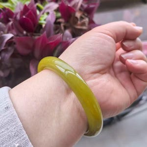 Könnte beinhalten: Ein glattes, olivgrünes Armreif-Armband. Das jadeähnliche Armband wird am Handgelenk getragen, mit einem verschwommenen Hintergrund aus lila Blumen. Das Armband hat eine glänzende Oberfläche und eine abgerundete Form.