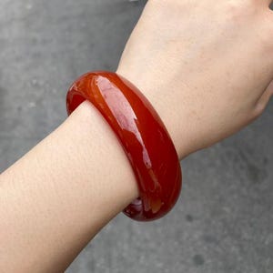 May include: A smooth, polished, solid red bangle bracelet with a rounded exterior profile and a glossy, translucent finish. The circular accessory features a deep, vibrant red hue throughout its entire structure, showcasing a consistent colour.