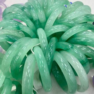 May include: A large collection of smooth, polished, translucent green jade-style bangles arranged in a circular, overlapping cluster. Each bangle features a rounded profile with varying shades of light to deep emerald green throughout.
