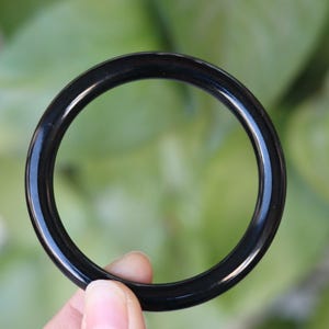 May include: A smooth, circular bangle bracelet with a polished, solid black finish. The bangle has a consistent, rounded profile and a glossy surface that reflects light, creating a clean, simple, and uniform appearance throughout the entire piece.