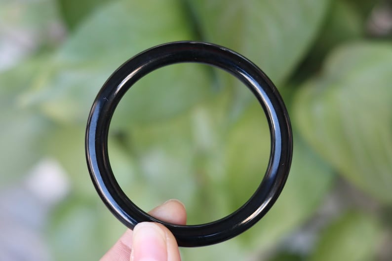 May include: A smooth, circular bangle bracelet with a polished, solid black finish. The bangle has a consistent, rounded profile and a glossy surface that reflects light, creating a clean, simple, and uniform appearance throughout the entire piece.