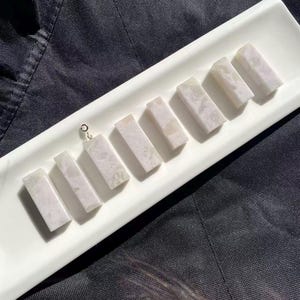 Puede incluir: Una colección de colgantes rectangulares de piedra clara, cada uno de aproximadamente 2,5 cm de largo, dispuestos sobre una bandeja rectangular blanca. Los colgantes tienen un aspecto veteado con sutiles variaciones de color. El fondo es una tela oscura y texturizada.