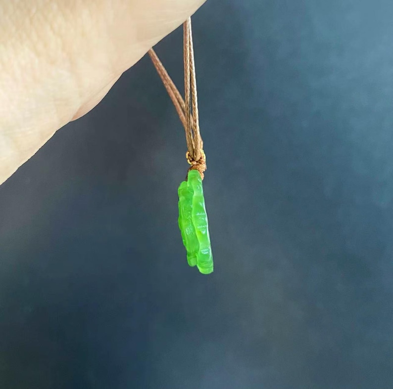 May include: A bright green pendant necklace with a floral design. The pendant is suspended from a brown cord. The background is a solid, dark blue-gray.