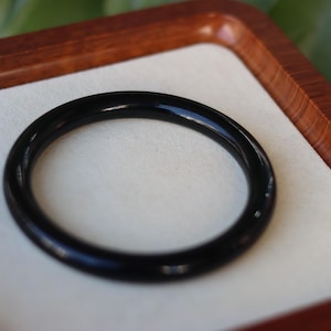 May include: A smooth, solid black circular bangle bracelet with a polished, reflective surface. The bangle has a uniform, rounded profile and a continuous, seamless design, presented as a single piece of jewelry with a deep, dark color.