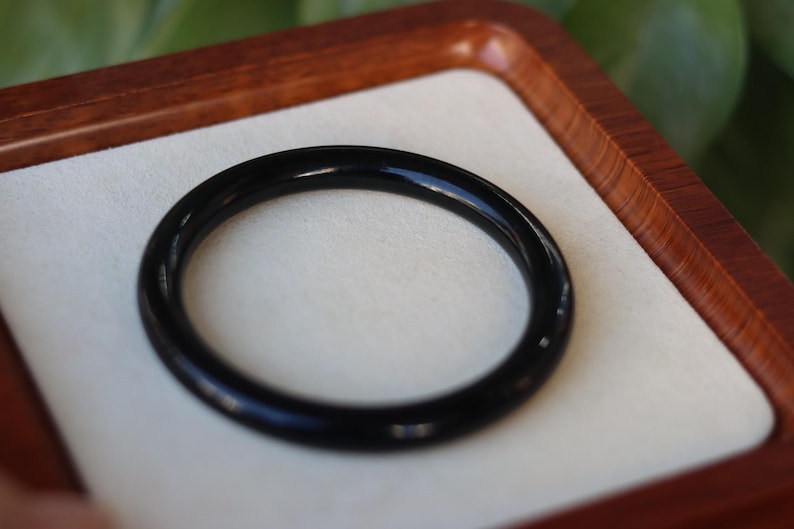 May include: A smooth, solid black circular bangle bracelet with a polished, reflective surface. The bangle has a uniform, rounded profile and a continuous, seamless design, presented as a single piece of jewelry with a deep, dark color.