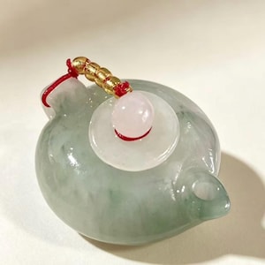 Könnte beinhalten: Ein kleines, rundes Teekannen-Ornament aus hellgrünem Jade mit einer glatten, polierten Oberfläche. Der Deckel ist mit einer blassrosa, kugelförmigen Perle und einer flachen weißen Scheibe verziert, die mit einer roten Schnur an kleinen Goldperlen befestigt ist.