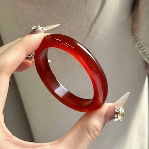Puede incluir: Un brazalete rojo translúcido y pulido. El brazalete liso y circular se sostiene sobre un fondo neutro. El color del brazalete es un rojo intenso y rico, con sutiles variaciones de tono. Una pieza elegante.
