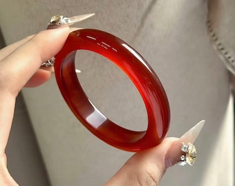 May include: A polished, translucent red bangle bracelet. The smooth, circular bracelet is held up against a neutral background. The color of the bracelet is a deep, rich red, with subtle variations in tone. This piece is a stylish accessory.