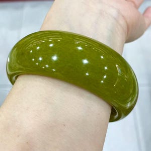 以下が含まれることがあります： 光沢のある磨き上げられた仕上げの、滑らかで無垢なオリーブグリーンのバングルブレスレット。円形のアクセサリーは、厚みのある丸みを帯びた形状で、光を反射して表面に繊細なハイライトを生み出す半透明の質感が特徴です。