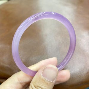 May include: A smooth, translucent lavender-colored bangle bracelet with a rounded profile. The circular jewelry piece features a consistent light purple hue throughout its solid, polished surface, reflecting light along its curved outer edge.