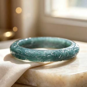 May include: A translucent, deep green jade bangle bracelet featuring intricate, hand-carved relief patterns of swirling clouds and dragon-like motifs encircling the entire outer surface of the polished, circular stone band.