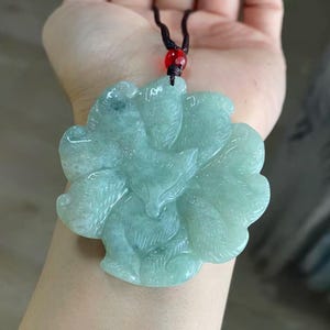 May include: A light green carved jade pendant featuring a nine-tailed fox design with intricate fur detailing. The pendant is suspended from a dark brown cord accented with a single small red bead at the top, showcasing a smooth polished finish.