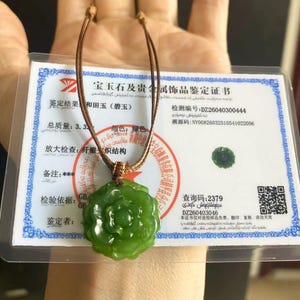 May include: A green jade pendant necklace with a flower design. The pendant is suspended from a brown cord. A certificate with text in a foreign language is visible in the background.
