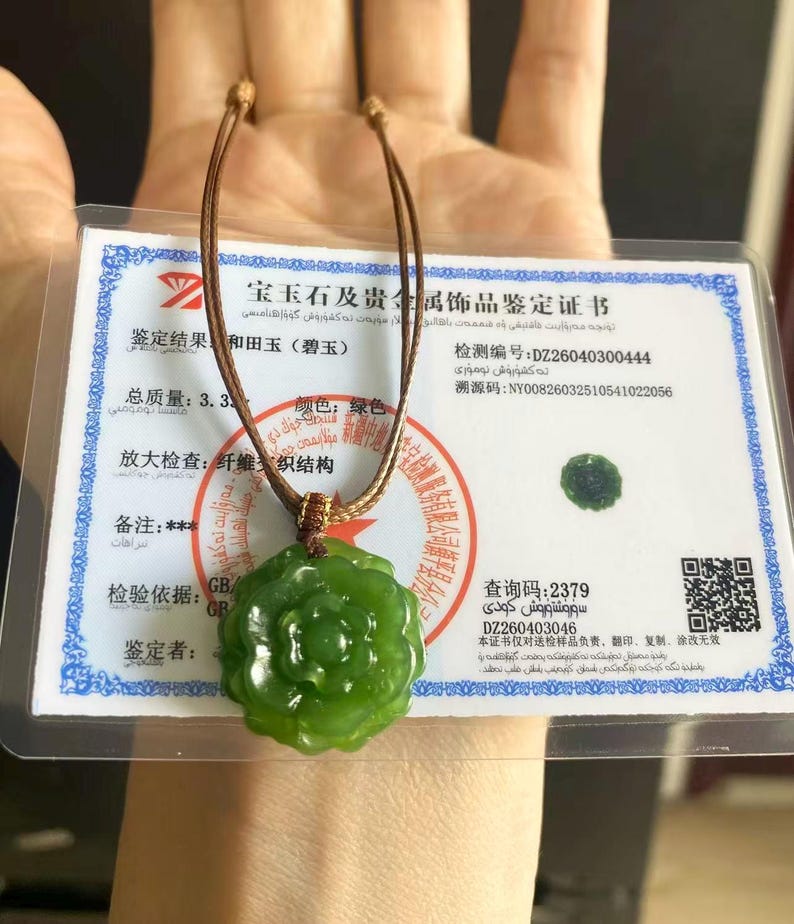May include: A green jade pendant necklace with a flower design. The pendant is suspended from a brown cord. A certificate with text in a foreign language is visible in the background.
