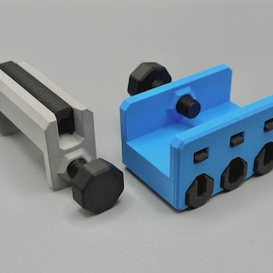 May include: Two 3D-printed clamps. One is light gray with a black insert and a black screw. The other is blue with black screws. The clamps are designed to hold objects in place.