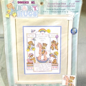 May include: A framed Dimensions Baby Hugs counted cross stitch kit. The design features teddy bears with umbrellas, a rocking horse, and the text "Kissed by the rain, warmed by the sun, a baby's life is full of fun!" The finished size is 9" x 12".