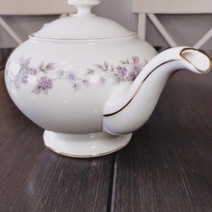 May include: A white porcelain teapot featuring a delicate floral pattern of small purple flowers and grey leaves encircling the body. The teapot has a matching lid with a small knob, a curved spout, and a gold-trimmed rim along the base and spout.