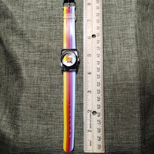 May include: A square-faced watch with a rainbow-striped band and a smiley face on the watch face. The watch band is approximately 9 inches long. The watch has a silver-colored metal case and a buckle clasp.