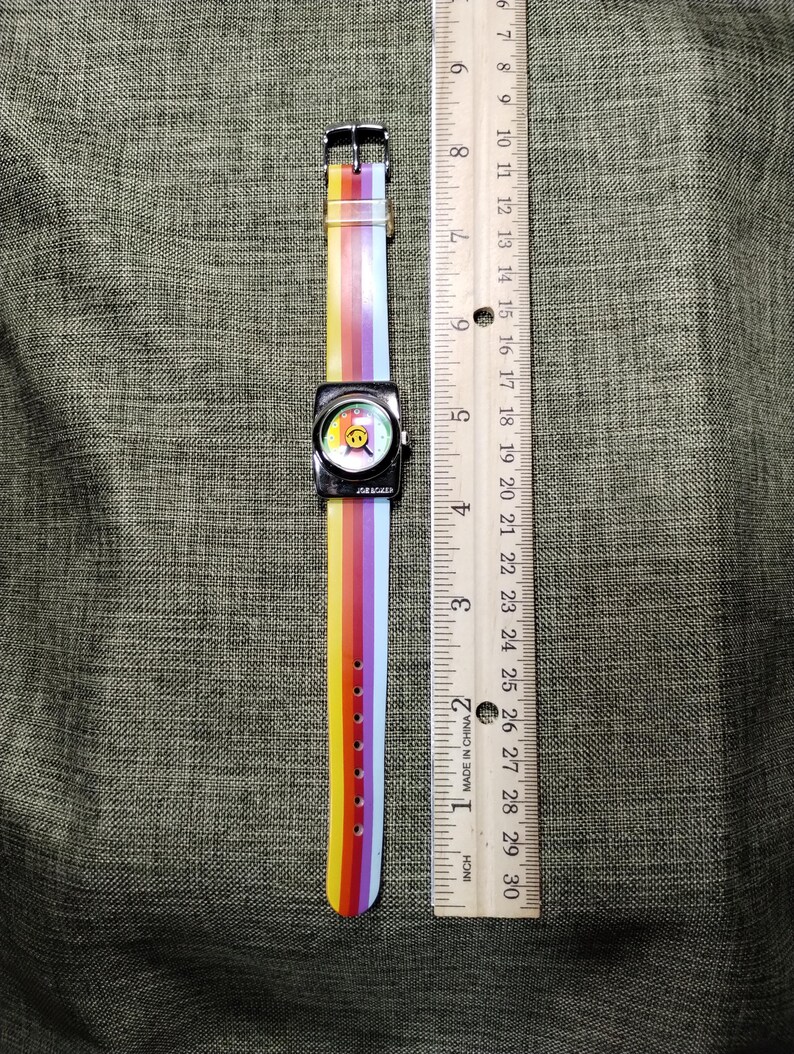 May include: A square-faced watch with a rainbow-striped band and a smiley face on the watch face. The watch band is approximately 9 inches long. The watch has a silver-colored metal case and a buckle clasp.