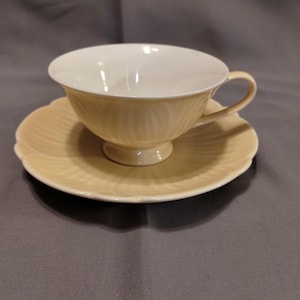 May include: A light yellow teacup and saucer set. The teacup has a white interior and a handle. The saucer is scalloped around the edge. The set is made of ceramic and is on a gray surface.