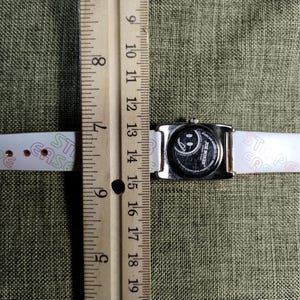 May include: A silver-toned rectangular watch with a white band. The watch face features a smiley face design. The band has the word "EASE" printed on it. The watch is measured with a wooden ruler.