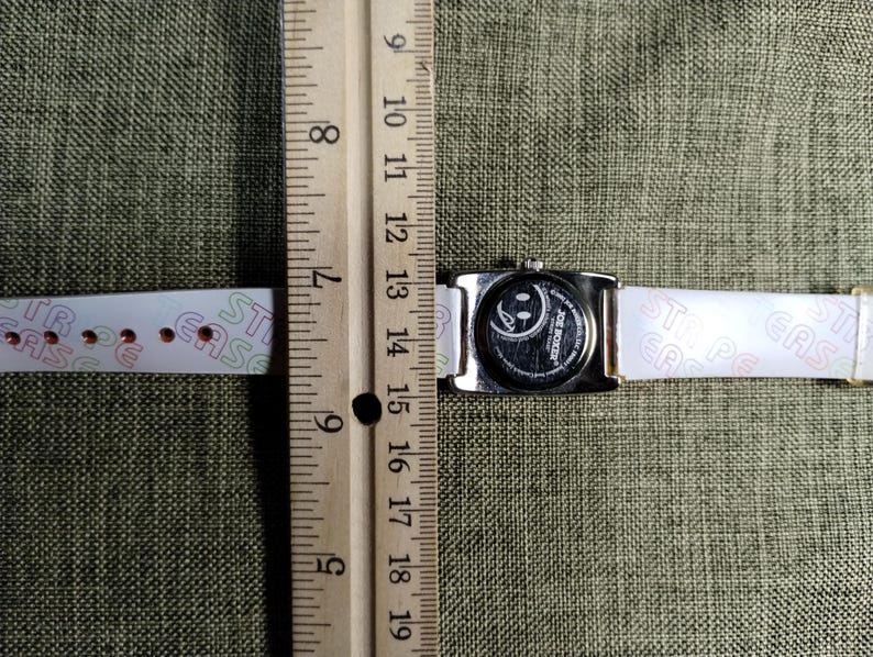 May include: A silver-toned rectangular watch with a white band. The watch face features a smiley face design. The band has the word "EASE" printed on it. The watch is measured with a wooden ruler.