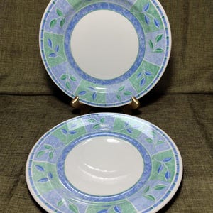 May include: Two white ceramic plates with a blue and green leaf design. The plates have a white center, a blue border, and a patterned outer rim with green and blue squares and leaf motifs. The plates are on a dark green surface.