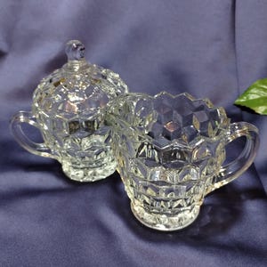 May include: Clear glass sugar bowl with lid and a creamer. Both pieces have a diamond-cut pattern and small handles. The sugar bowl has a small knob on the lid. The creamer has a scalloped rim. The set is on a dark blue background.