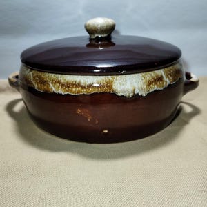 May include: A brown ceramic casserole dish with a lid. The dish has two handles and a white and brown rim. The lid has a small, round knob. The dish is on a tan surface.