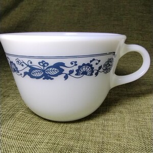 May include: A white milk glass cup featuring a rounded bowl shape and a curved handle. The exterior is decorated with a horizontal band of dark blue floral and vine patterns, including stylized leaves and blossoms encircling the upper section.