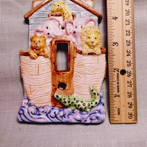 May include: A ceramic light switch plate shaped like Noah's Ark, featuring a giraffe, two elephants, a bear, a lion, and a crocodile. The plate is positioned next to a wooden ruler, showing a height of approximately 5 inches for the object.
