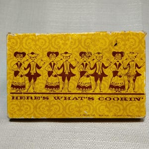 May include: A rectangular yellow box featuring a repeating damask pattern and a row of brown illustrations depicting four pairs of figures in historical attire holding cooking utensils. The text reads: HERE'S WHAT'S COOKIN' and CURRENT, INC. BOX 2020.