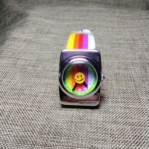 May include: A square, silver-toned wristwatch with a colorful smiley face on the dial. The watch face features a rainbow design and a yellow smiley face. The watch band is a rainbow of colors. The watch is a fun, retro accessory.