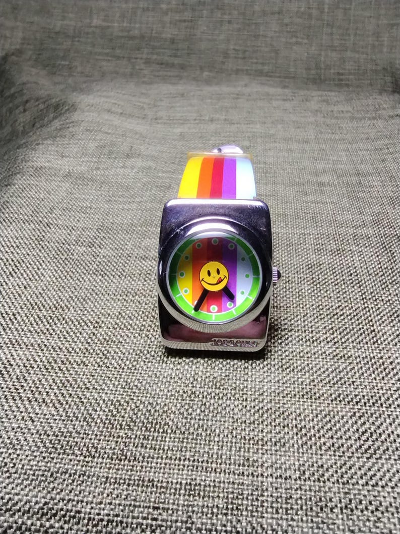 May include: A square, silver-toned wristwatch with a colorful smiley face on the dial. The watch face features a rainbow design and a yellow smiley face. The watch band is a rainbow of colors. The watch is a fun, retro accessory.