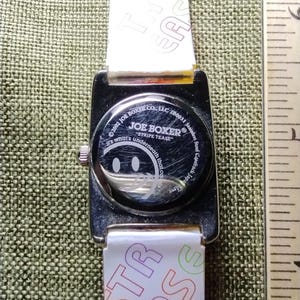 May include: A Joe Boxer wristwatch with a white strap featuring the text "STRIPE TEASE" and "TYPE EASE". The watch face has a smiley face design. The watch case is stainless steel.