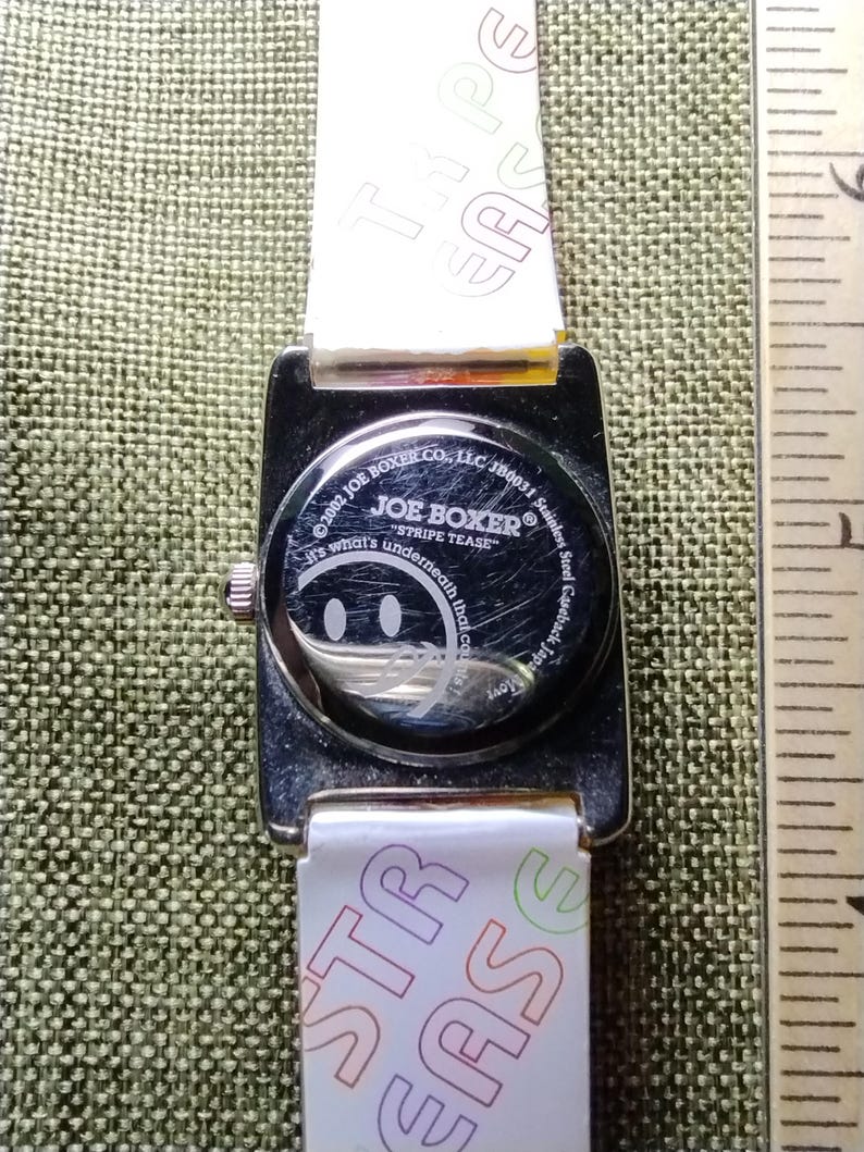 May include: A Joe Boxer wristwatch with a white strap featuring the text "STRIPE TEASE" and "TYPE EASE". The watch face has a smiley face design. The watch case is stainless steel.
