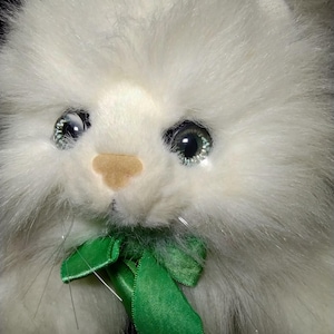 May include: A plush toy cat with long, fluffy white fur, large reflective green eyes, and a small tan nose. The stuffed animal features thin, clear plastic whiskers and a bright green satin ribbon tied in a bow around its neck area.