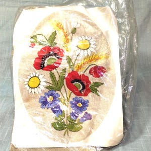 May include: An oval-shaped needlepoint kit featuring a floral bouquet of red poppies, white daisies, blue flowers, and wheat stalks. The embroidery is on a beige fabric background. The kit is sealed in a clear plastic bag.